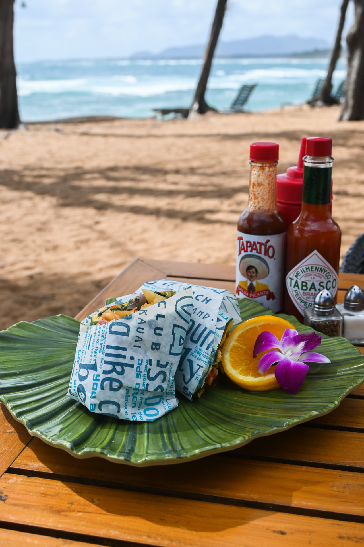 Breakfast wrap on a ti leaf plate with orange and orchid, Kaua'i shoreline behind
