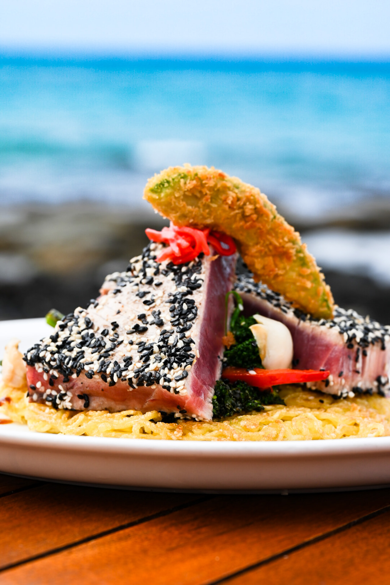 Sesame-crusted seared ahi over crispy noodles with tempura avocado, ocean behind